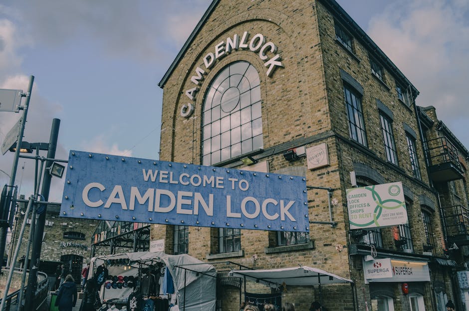An exterior view of the Camden Lock building showcasing its brick facade and large arched window. A prominent blue sign reading 'Welcome to Camden Lock' is hung below a sign with the city name, indica