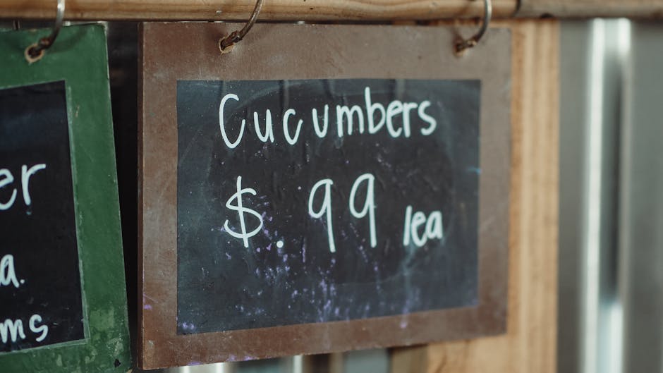 A close-up of a black chalkboard sign with a brown wooden frame, displaying the handwritten message 'Cucumbers $.99 ea' in white chalk. The sign is attached to a wooden surface with metal hooks, indic