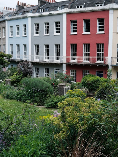 A panoramic view of a city skyline seen from a small, terraced garden surrounded by lush greenery. In the foreground, well-maintained grass alongside garden beds with low shrubs and leaf litter are vi
